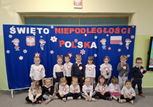 Dzieci z grupy 1 ubrane na galowo pozują do zdjęcia na tle dekoracji na polskich barwach.