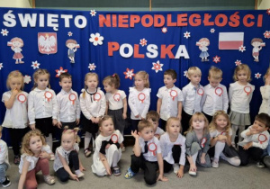 Dzieci z grupy 2 ubrane na galowo pozują do zdjęcia na tle dekoracji na polskich barwach.