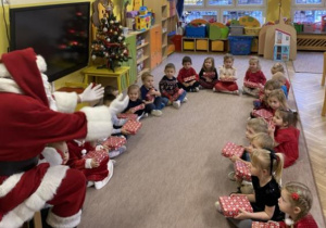 Dzieci z grupy 2 siedzą w kole na dywanie wraz z Mikołajem i śpiewają mu piosenkę.