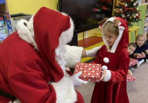 Mikołaj wręcza prezent dziewczynce w czerwonej sukience.